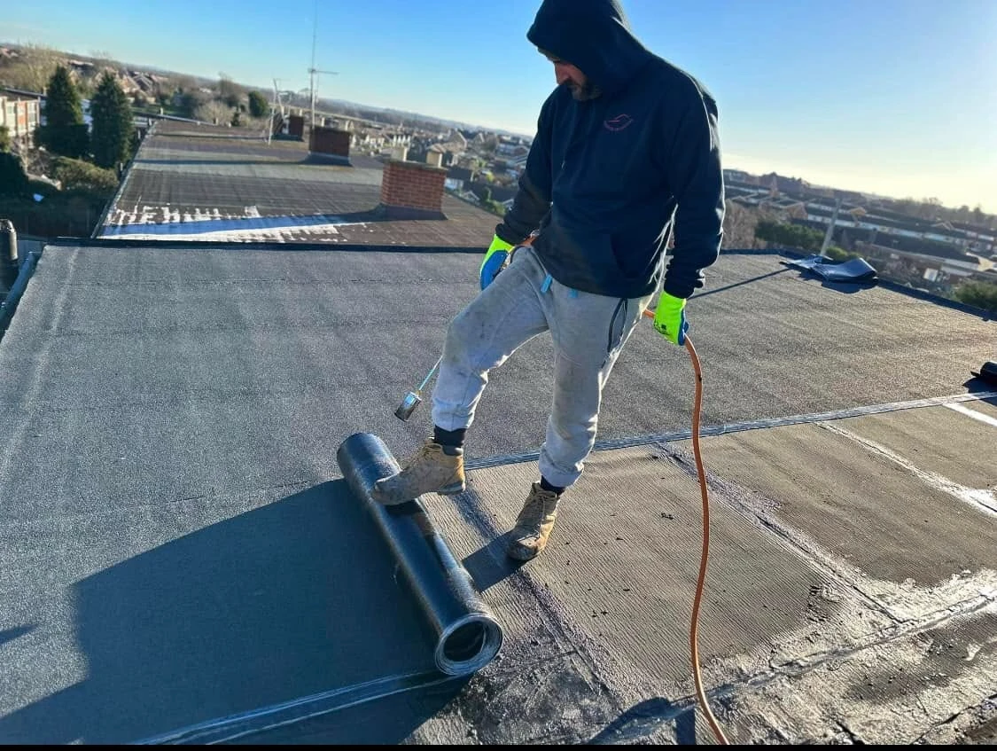 Worker wearing a hooded sweatshirt, gloves, and boots applying roofing felt on a flat rooftop using a propane torch and foot to unroll the material on a sunny day.
