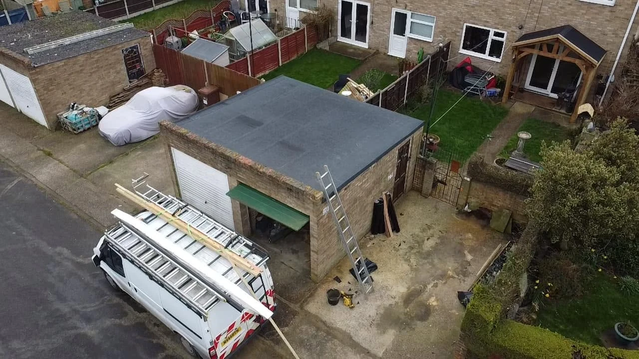 Aerial view of a residential garage with an open door, a ladder leaning on the wall, a white van with ladders on top, a car covered with a white sheet, and neighboring garden areas with grass, shrubs, and patio furniture.