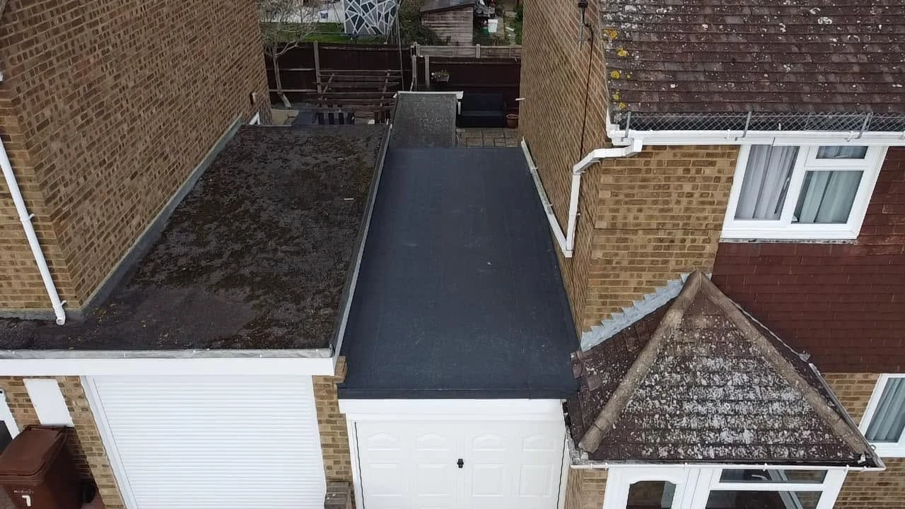 Top view of attached garages between two brick houses, one with a mossy flat roof and the other a dark smooth flat roof, with a sloped tiled roof porch and white framed windows.