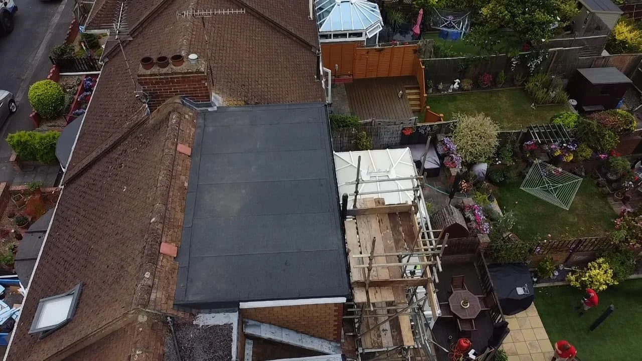 Aerial view of a house with a brown tiled roof and a flat black roof section, adjacent to a garden with green lawn, patio furniture, scaffolding, plants, and two people in red shirts.