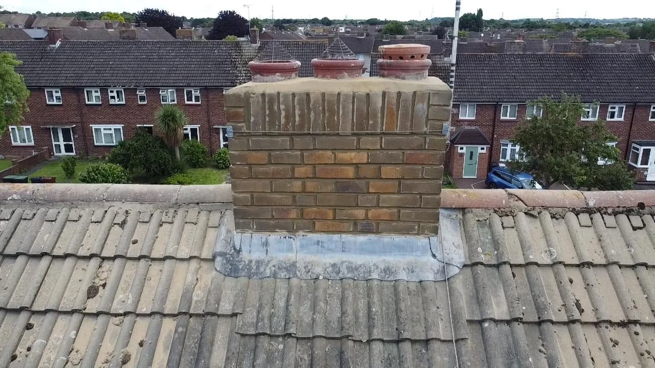 Brick chimney with three clay chimney pots on a sloped, tiled rooftop, with residential houses and trees visible in the background under a clear sky.