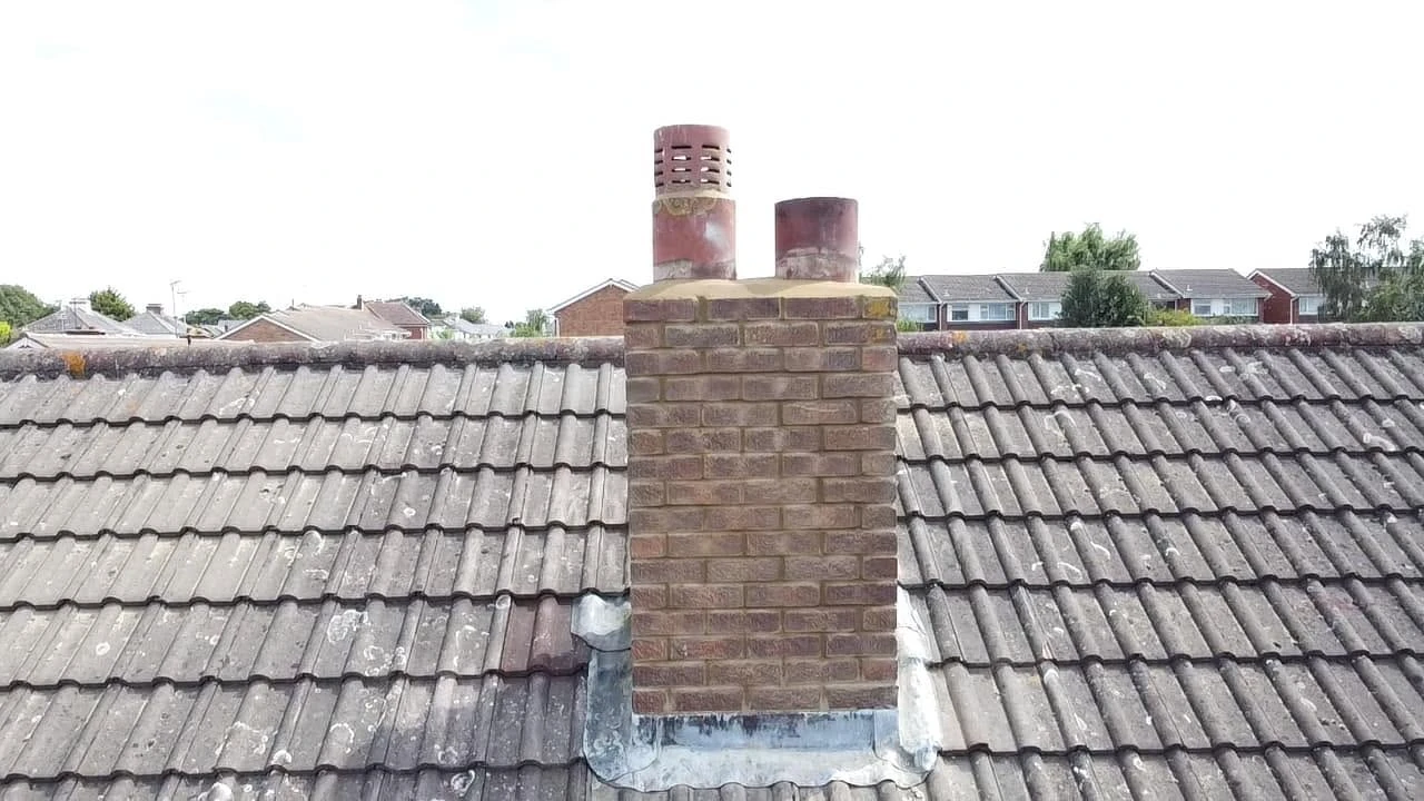Brick chimney with two metal flues on a weathered tiled roof with houses and trees in the background under a clear sky.