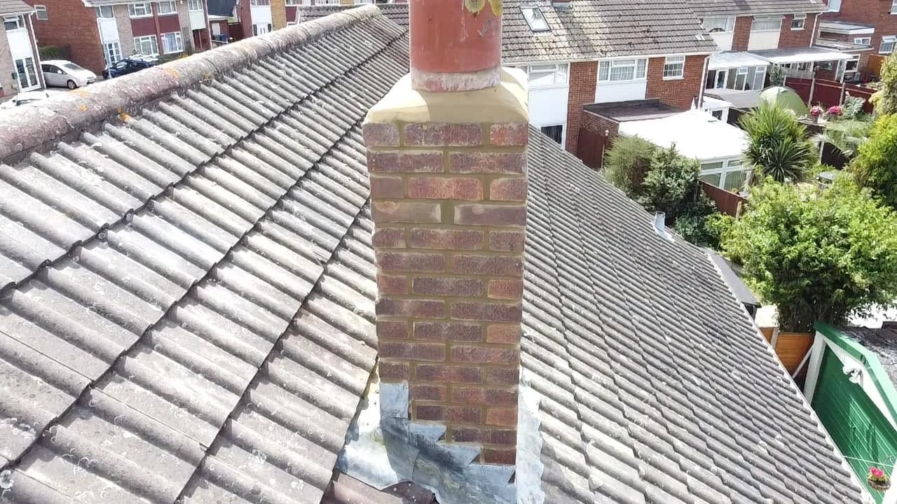 Brick chimney on a ridged gray tiled roof of a residential house with neighboring homes in the background.