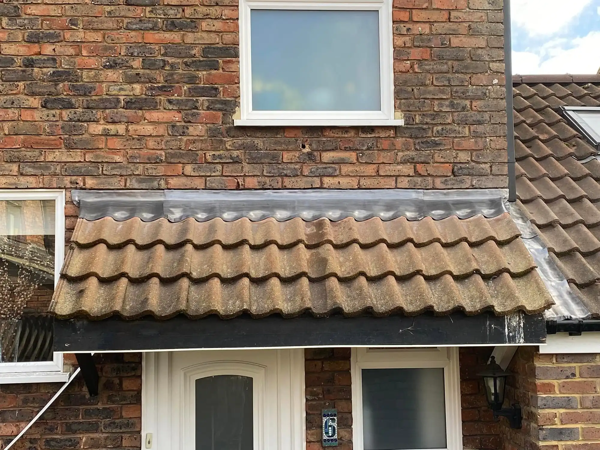 Front entrance of a brick house with a white door and window topped by a small tiled roof canopy with a metal flashing above it.