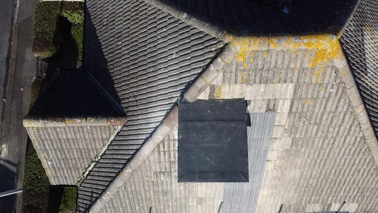 Overhead view of a large rooftop with gray tiles and a smaller black rectangular structure near the center, with some yellow moss or lichen on parts of the roof tiles.