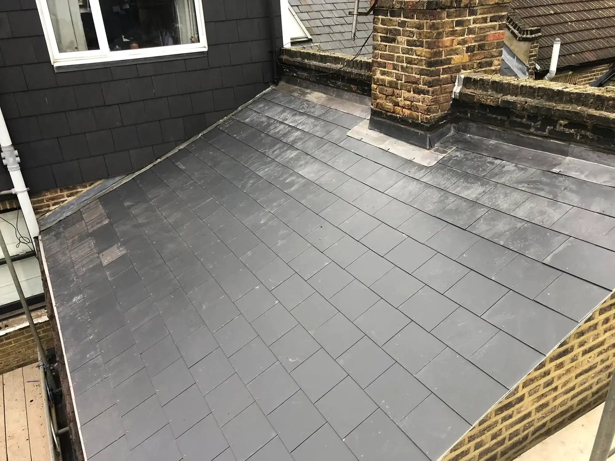 Sloped roof covered with rectangular gray tiles next to a brick chimney and dark shingle wall with a window.