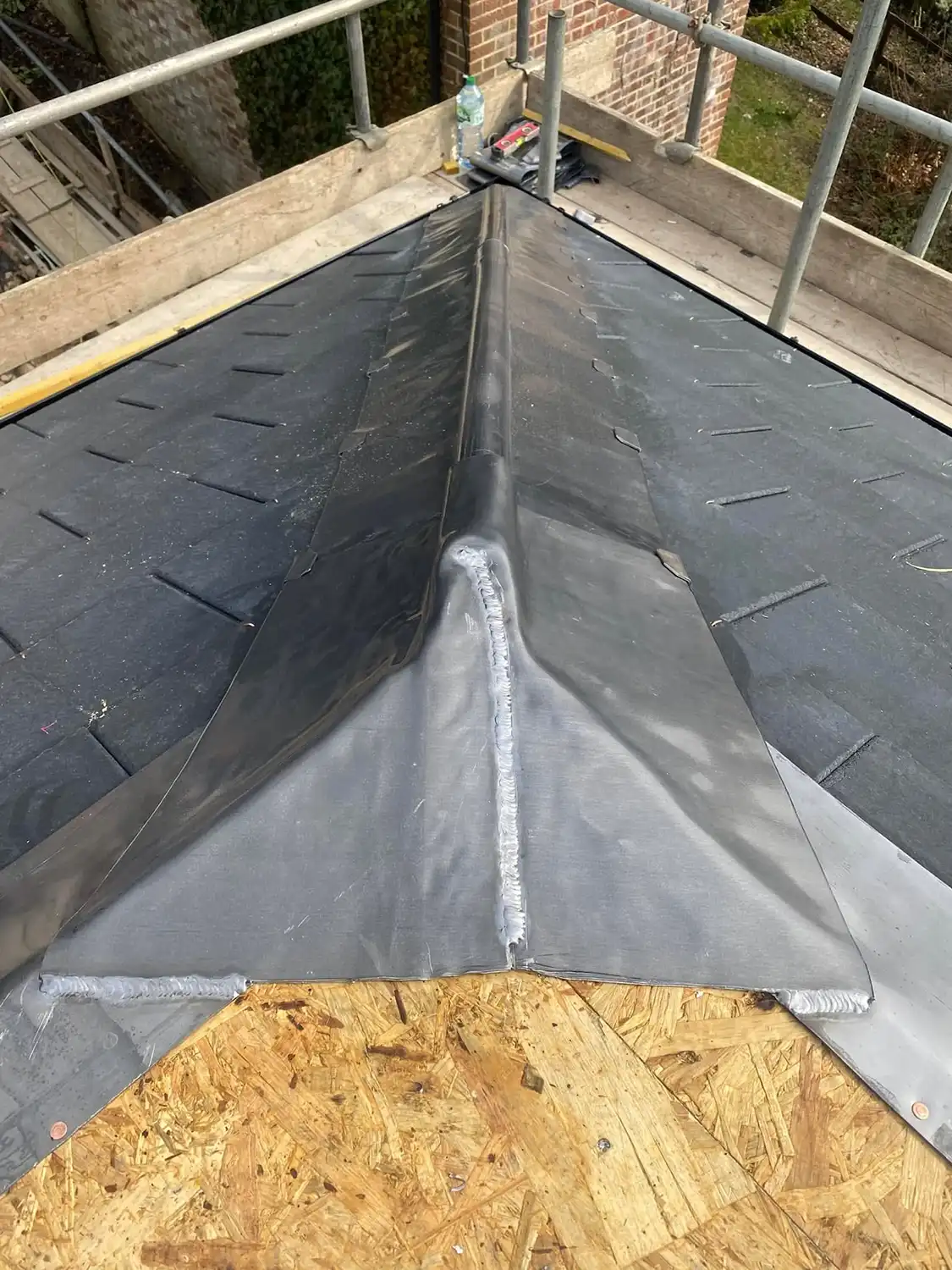 Close-up of a roof's ridge with black waterproof membrane installed, showing a seam sealed with thick welding material, wooden board at the base, and scaffolding around the edge.