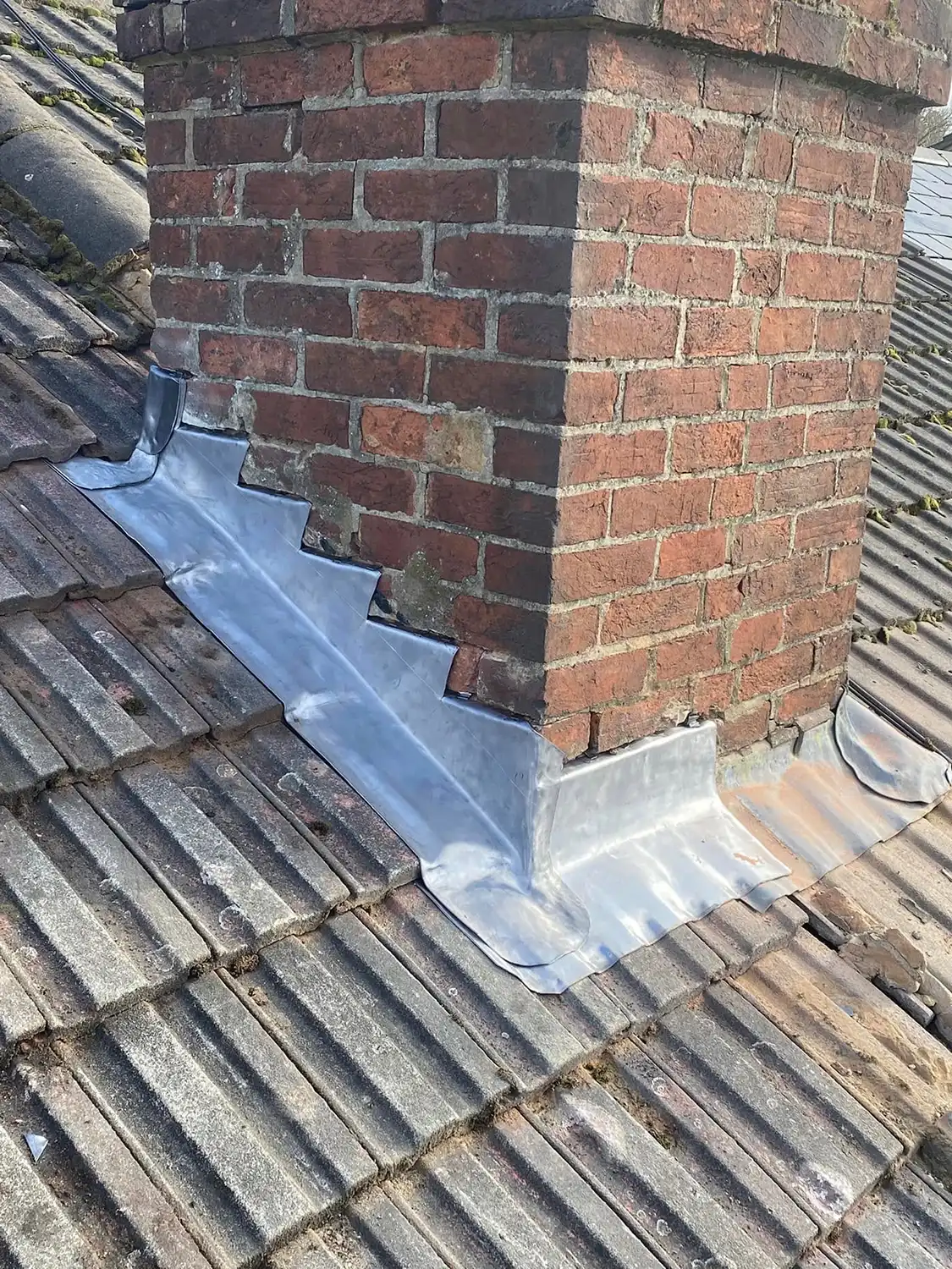 Red brick chimney on a tiled roof with new silver metal flashing installed around its base to prevent water leakage.