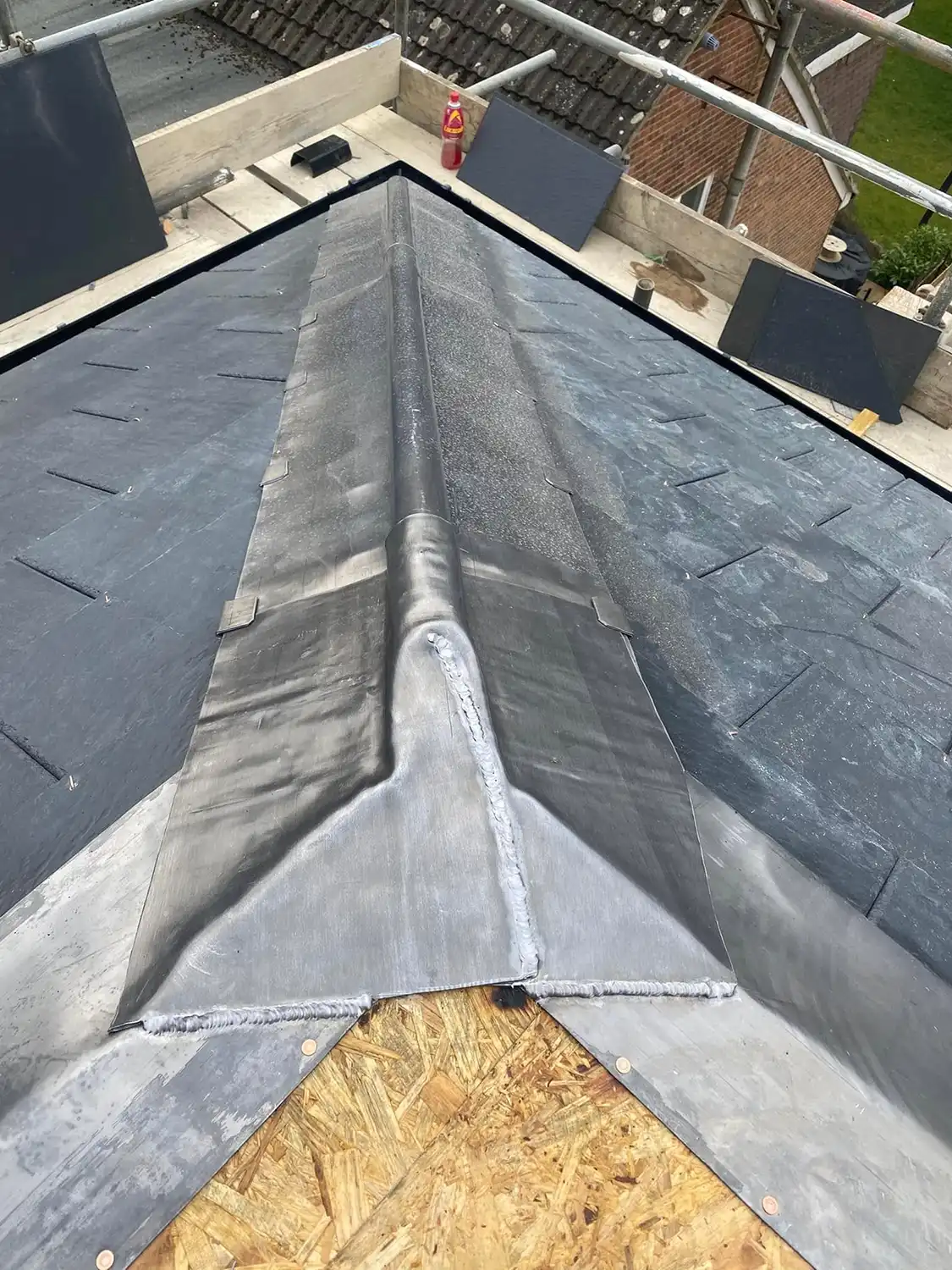 Close-up of a roof ridge with newly installed lead flashing and dark slate tiles on both sides, viewed from above with scaffolding and houses in the background.