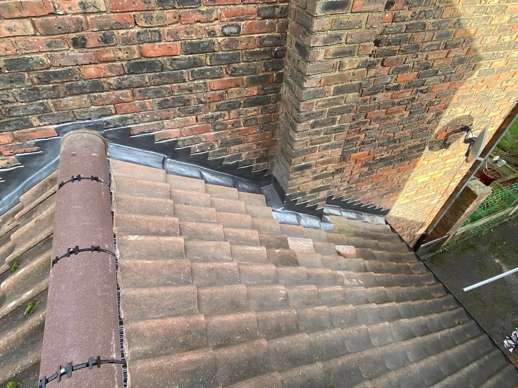 Section of pitched roof with brown tiles and ridge capped with secured tiles, adjacent to brick walls sealed with stepped lead flashing.