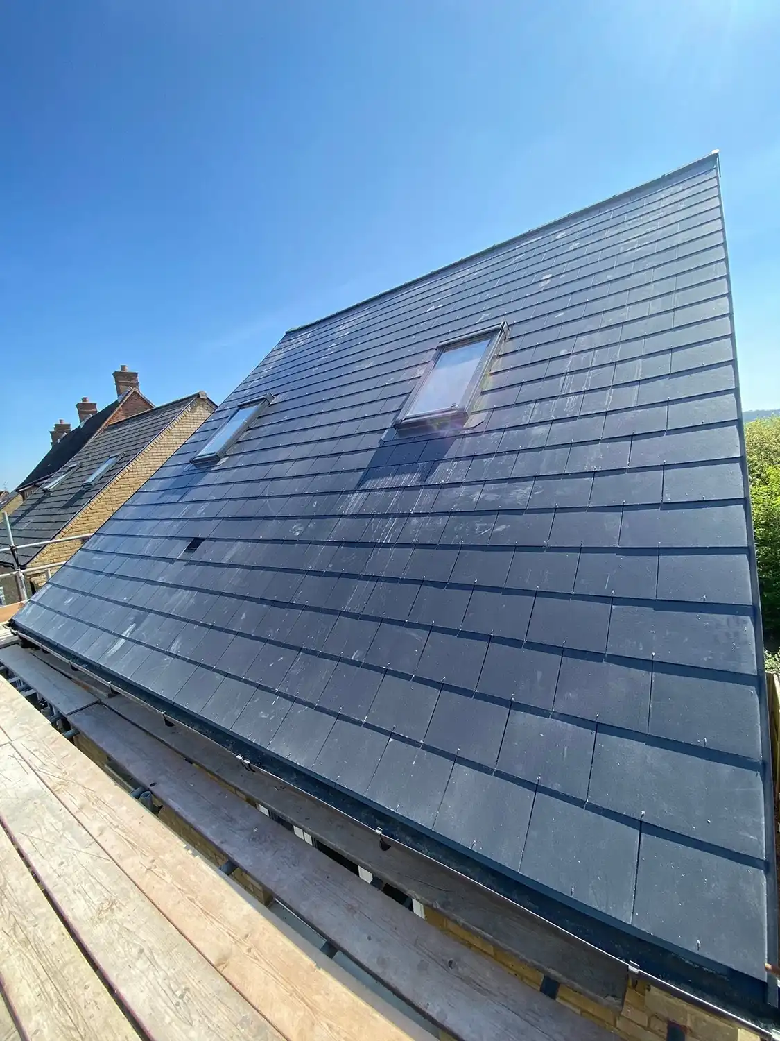 A freshly tiled dark roof with two skylights, viewed from scaffolding under a clear blue sky with neighboring rooftops visible on the left.