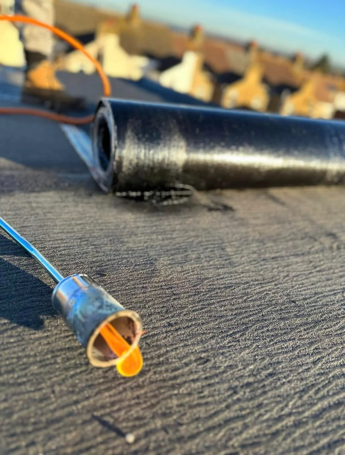 Rolled black roofing membrane on a textured roof surface with a metal and orange-handled tool resting beside it under clear blue sky.