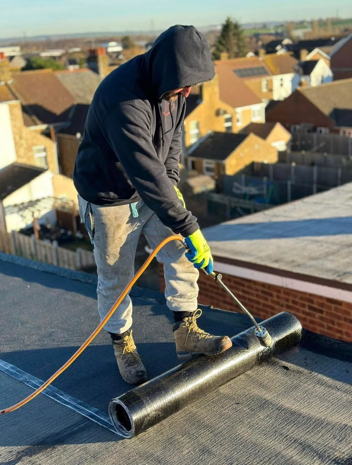 Worker wearing gloves, hoodie, and boots applying roofing material with a torch on a flat roof under clear sky in a residential neighborhood.