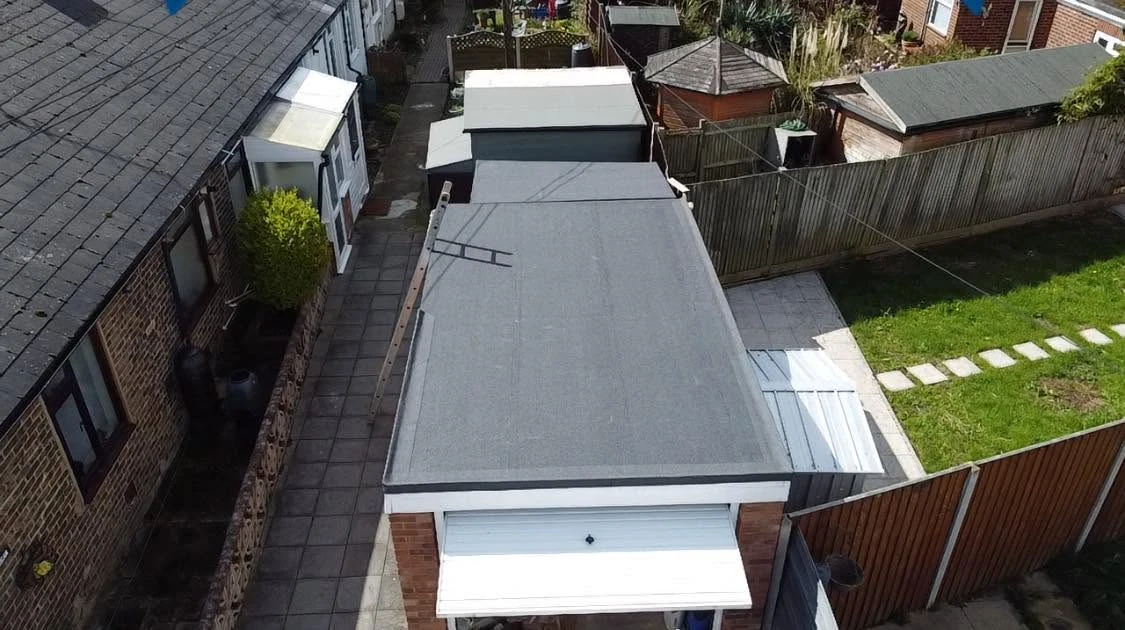 Aerial view of a row of terraced houses with flat grey roofs, a paved path beside them, a small garden with a lawn and stepping stones, and wooden fences separating properties.