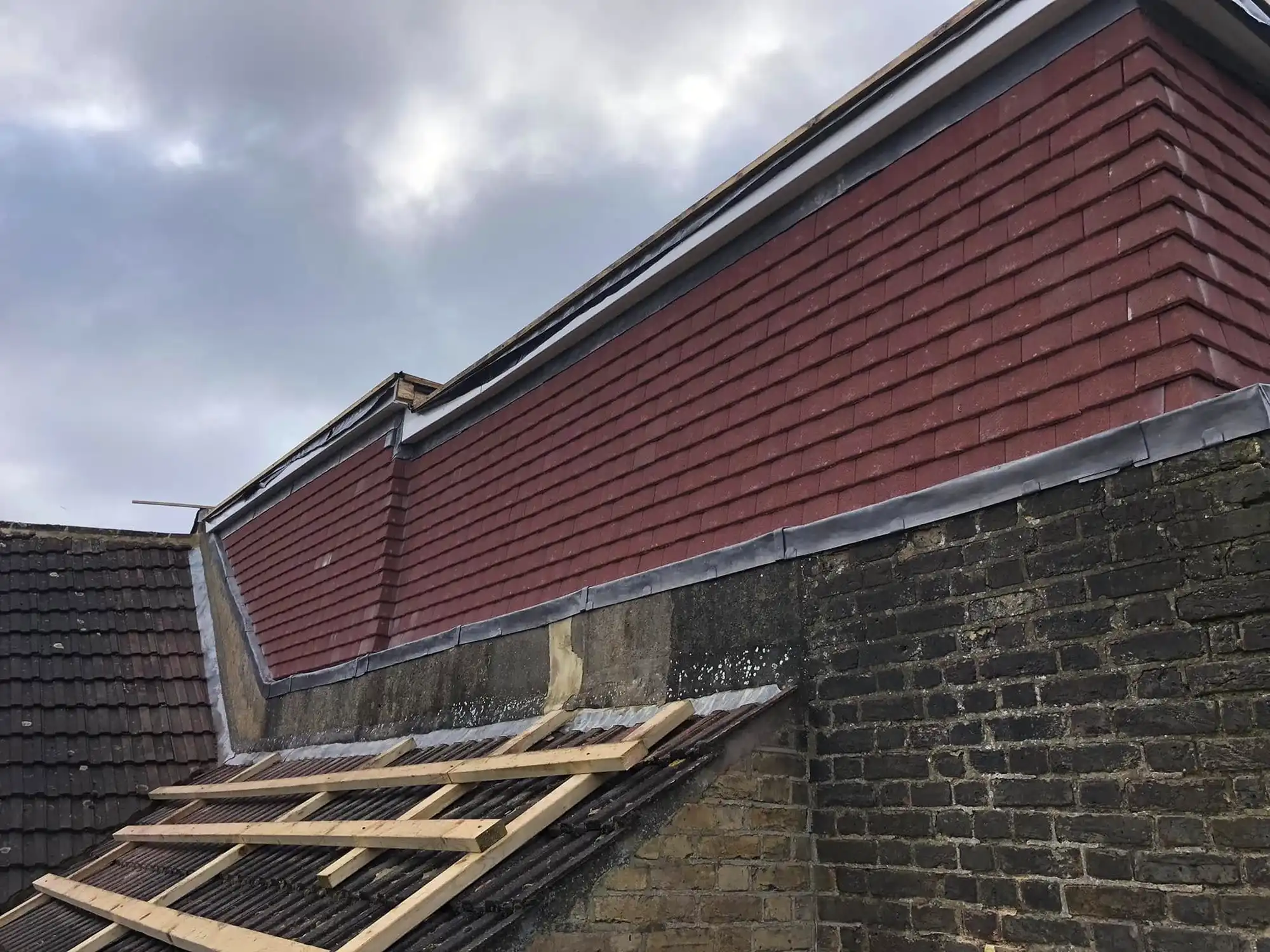 Exterior view of a building's roof area showing a section with brown shingles, dark tiled roof with wooden battens, and exposed brickwork wall under a cloudy sky.