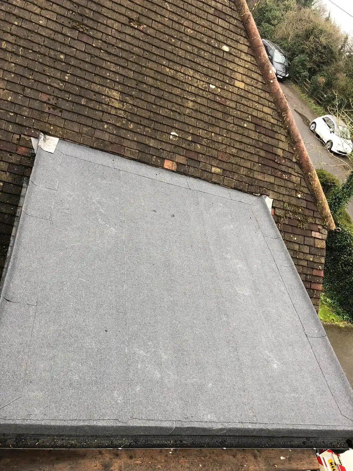 View of a flat roof covered with a smooth gray membrane next to a sloped roof with brown shingles, with parked cars and greenery visible in the background.