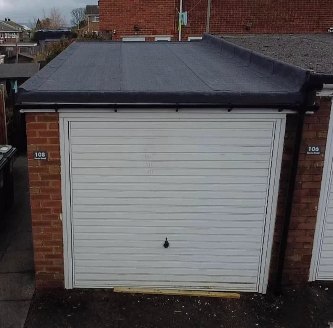 Front view of a closed white garage door with horizontal panels, flanked by brick walls with house numbers 108 on the left and 106 on the right, and a flat dark roof on top.